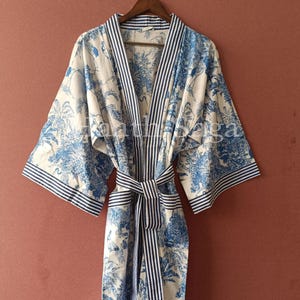 Könnte beinhalten: Blau-weiß gemusterter Bademantel mit gestreiftem Gürtel und Besatz. Der Bademantel hat ein blaues botanisches Design auf weißem Hintergrund. Der Bademantel hängt an einem Holzbügel.