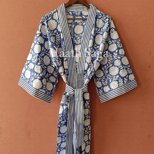 May include: A blue and white floral print robe with a striped trim and matching belt. The robe is hanging on a wooden hanger against a solid brown background. The floral pattern is a repeating design.