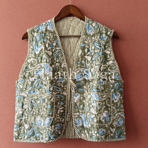 Puede incluir: Un chaleco acolchado sin mangas con un estampado floral en tonos verdes, azules y blancos. El chaleco tiene dos bolsillos delanteros y un forro a rayas. Cuelga de una percha de madera sobre un fondo liso.