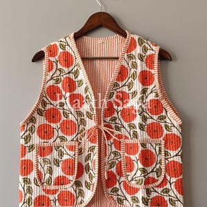 Pode incluir: Um colete acolchoado com padrão floral. O colete apresenta um fundo branco com grandes flores laranja e folhas verdes. Possui dois bolsos frontais e um fecho de laço. A borda e o forro têm um padrão listrado.