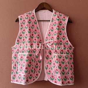 Puede incluir: Un chaleco acolchado rosa con un estampado floral de flores rojas y hojas verdes. El chaleco tiene dos bolsillos y un cierre de lazo. Cuelga de una percha de madera sobre un fondo marrón.