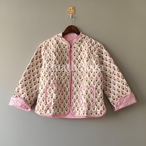 May include: A quilted jacket with a floral pattern in pink and green on a white background. The jacket has a round collar, two pockets, and pink striped trim. The jacket is hanging on a wooden hanger.