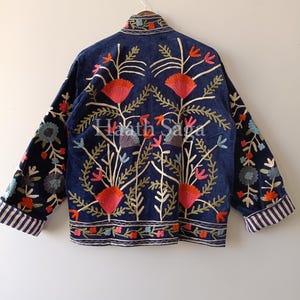 May include: A navy blue jacket with intricate floral embroidery in red, orange, green, and blue. The jacket features a relaxed fit, long sleeves, and striped cuffs. The back of the jacket displays the text "Haath Saga".