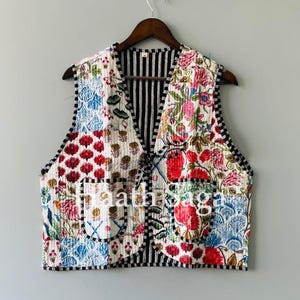 Könnte beinhalten: Eine gesteppte Weste mit Patchwork-Design mit floralen und geometrischen Mustern in Rot, Blau und Weiß. Die Weste hat ein schwarz-weiß gestreiftes Innenfutter und schwarze Paspeln. Sie hängt an einem Holzkleiderbügel.