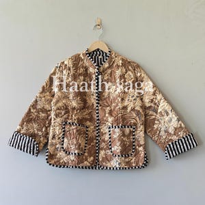 May include: A quilted jacket with a floral pattern in shades of brown and cream. The jacket has black and white striped trim on the cuffs, pockets, and edges. It is hanging on a wooden hanger against a neutral wall. The text "Haath saga" is visible on the jacket.