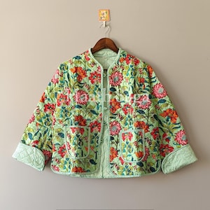 May include: A quilted jacket with a floral pattern in red, orange, and pink on a light green background. The jacket has two pockets and striped trim on the cuffs and edges. It is hanging on a wooden hanger.