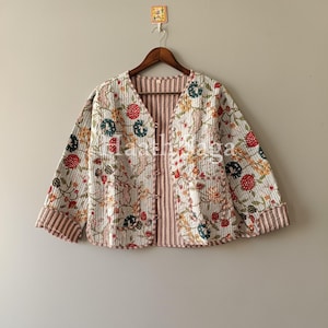 May include: A quilted jacket with a floral pattern in red, blue, and yellow on a white background. The jacket has a V-neck, long sleeves, and a striped lining. It is hanging on a wooden hanger.