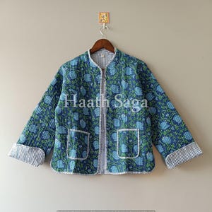 Könnte beinhalten: Gesteppte Jacke mit blau-grünem Blumenmuster. Sie hat einen Reißverschluss, zwei Taschen und gestreifte Manschetten. Der Text "Haath Saga" ist auf der Vorderseite aufgedruckt.