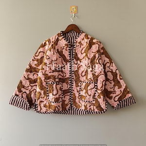 Könnte beinhalten: Wendejacke mit rosa-braunem Tiermuster. Die Jacke hat einen schwarz-weiß gestreiften Besatz an den Manschetten, Taschen und am Kragen. Sie hängt an einem Holzbügel vor neutralem Hintergrund.
