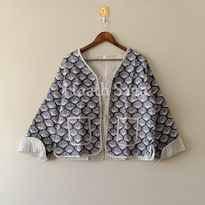 Könnte beinhalten: Eine marineblaue Jacke mit weißem Muschelmuster und zwei Vordertaschen hängt an einem Holzbügel. Das Innere und die Manschetten der Jacke weisen ein weißes und marineblaues Streifendesign auf. Der Text "Haath Saga" ist sichtbar.