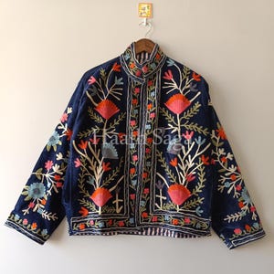 May include: A navy blue jacket with intricate floral embroidery in shades of orange, pink, green, and blue. The jacket features a high collar and is hanging on a wooden hanger. The text "Haar Saga" is visible on the jacket.