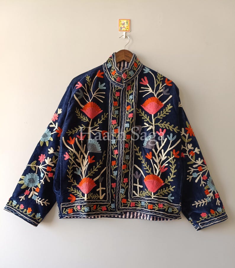 May include: A navy blue jacket with intricate floral embroidery in shades of orange, pink, green, and blue. The jacket features a high collar and is hanging on a wooden hanger. The text "Haar Saga" is visible on the jacket.