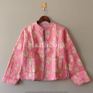 Könnte beinhalten: Rosa gesteppte Jacke mit goldenem Blumenmuster und zwei Vordertaschen. Die Jacke hat einen Stehkragen und einen durchgehenden Reißverschluss. Der Text "Haath Saga" ist auf der Vorderseite aufgedruckt.