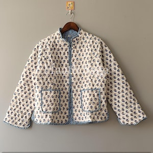 Puede incluir: Chaqueta acolchada color crema con un estampado floral azul. La chaqueta tiene cuello mandarín, dos bolsillos delanteros y una cremallera completa. Los bordes están ribeteados con tela azul. La chaqueta está colgada en una percha de madera.