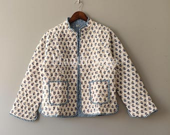 May include: Cream-colored quilted jacket with a blue floral pattern. The jacket has a mandarin collar, two front pockets, and a full-length zipper. The edges are trimmed with blue fabric. The jacket is hanging on a wooden hanger.