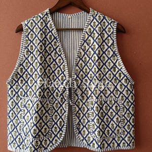 Può includere: Un gilet senza maniche con un motivo a rombi blu navy e verde oliva, con interno e bordi a righe. Il gilet è appeso a una gruccia di legno su uno sfondo marrone.