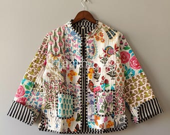 May include: A colourful quilted jacket with a floral and butterfly design. The jacket displays a mix of colours, including pink, blue, and green, with black and white striped accents on the cuffs and trim. It has a mandarin collar and a black zipper.