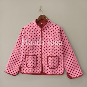 Könnte beinhalten: Eine hellrosa gesteppte Jacke mit dunkelrotem Punktemuster, Stehkragen, langen Ärmeln und zwei aufgesetzten Vordertaschen. Die Jacke hat einen dunkelroten Saum an den Kanten und eine durchgehende Knopfleiste mit dem Text Haath saga.