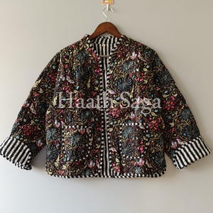 以下が含まれることがあります： ピンク、青、黄色で花柄の黒いキルティングジャケット。袖口と襟には白と黒のストライプのトリムが付いています。「Haath Saga」の文字が前面に見えます。