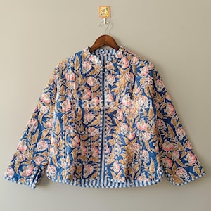 Peut inclure: Veste matelassée à motifs floraux bleus, roses et bruns. La veste est dotée d'une fermeture éclair, de deux poches et d'une bordure rayée. Elle est suspendue à un cintre en bois.
