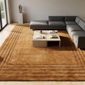 Könnte beinhalten: Ein großer, rechteckiger, brauner Teppich mit einem geometrischen Bordürenmuster. Der Teppich befindet sich in einem modernen Wohnzimmer, mit einem grauen Ecksofa, einem Couchtisch aus hellem Holz und einer Topfpflanze. Der Raum hat große Fenster und Holzböden.