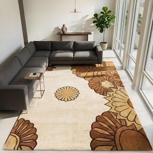 May include: A large, rectangular area rug with a floral design in shades of brown and tan. The rug features a cream-colored background with large, stylized flower patterns along the edges and a single flower in the center. The rug is placed in a living room setting.
