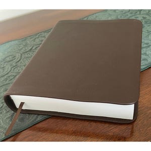 May include: A brown leather-bound book with a matching bookmark. The book is closed, with the pages visible along the edge. The book is resting on a patterned green and brown surface.