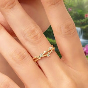 May include: A gold ring featuring a twig design, embellished with small, clear gemstones. The ring is displayed on a finger, highlighting its intricate details. The background shows a tropical scene with vibrant greenery and flowers.