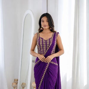 May include: A deep purple sleeveless blouse and matching draped skirt featuring intricate gold embroidery along the neckline, bodice, and hem. The ensemble includes a long, flowing purple fabric panel that hangs from the shoulder to the floor.