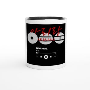 May include: A white ceramic mug with a black interior and rim. The mug features the Korean word "Arirang" in red, along with three white circular designs. Below, the text "NORMAL" and "BTS" are displayed, with a music player interface.