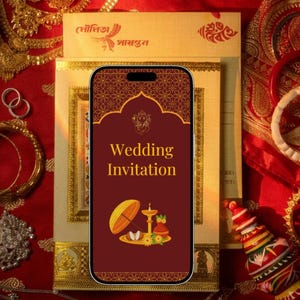 May include: A smartphone displays a wedding invitation with a maroon background and gold accents. The image is surrounded by gold bangles, rings, and traditional Indian wedding accessories on a red fabric.