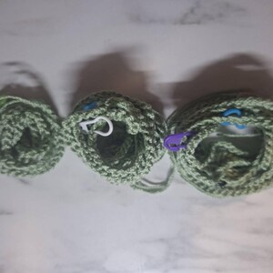 May include: Three crocheted items in varying sizes, each in a light green hue. The items appear to be circular, with visible stitch patterns. Stitch markers in white, blue, and purple are attached to the crocheted pieces. The items are arranged on a light-colored surface.