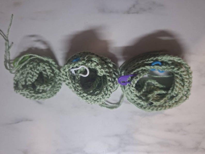 May include: Three crocheted items in varying sizes, each in a light green hue. The items appear to be circular, with visible stitch patterns. Stitch markers in white, blue, and purple are attached to the crocheted pieces. The items are arranged on a light-colored surface.