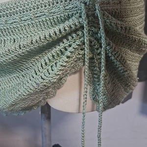 May include: A sage green crochet top with adjustable straps. The top features a scalloped edge and intricate stitch patterns. The straps are made of the same yarn and are tied at the top. The top is displayed on a mannequin.