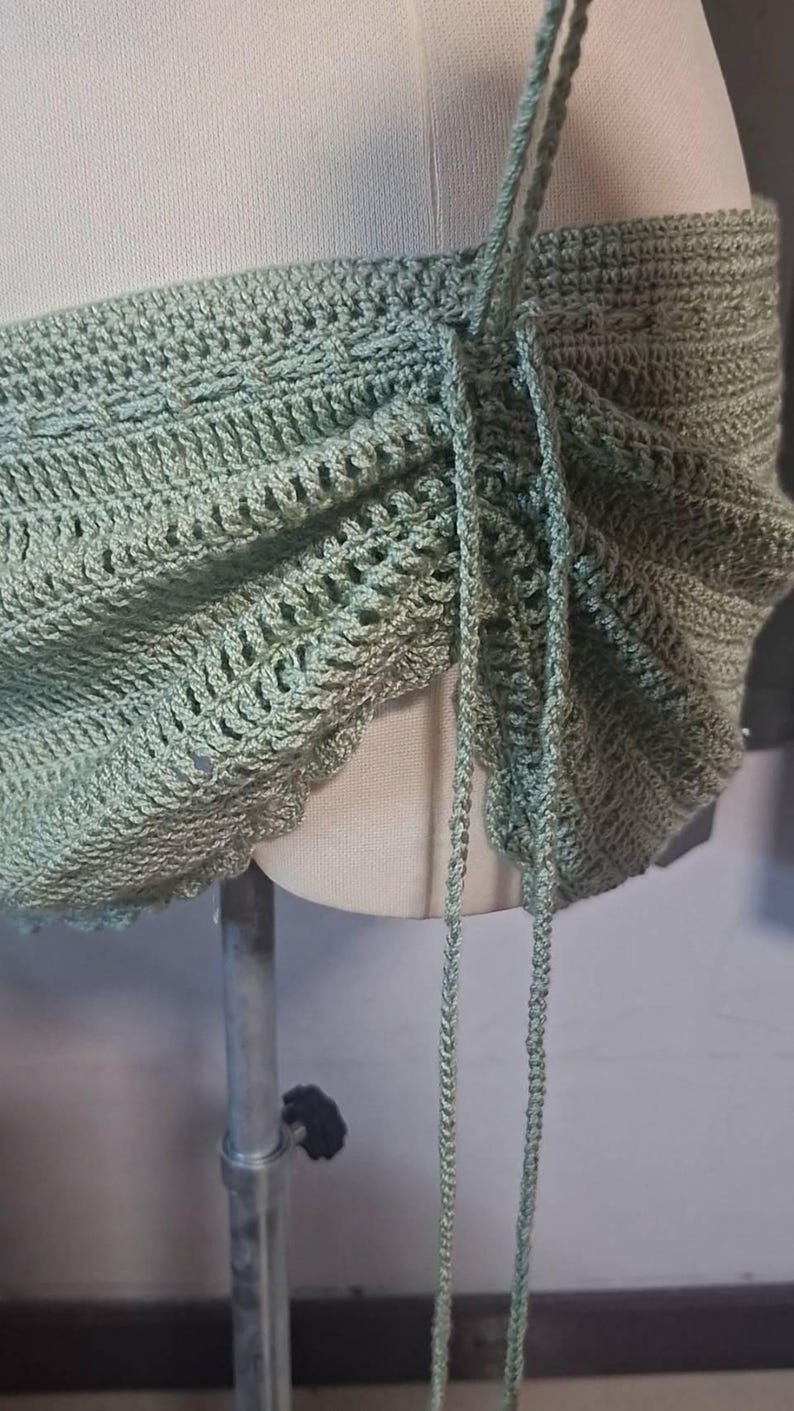 May include: A sage green crochet top with adjustable straps. The top features a scalloped edge and intricate stitch patterns. The straps are made of the same yarn and are tied at the top. The top is displayed on a mannequin.