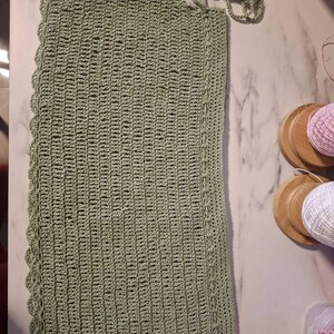 May include: A sage green, rectangular crochet piece with a scalloped edge. The handmade item features a textured stitch pattern and a decorative cord with wooden beads. The piece is likely a handmade home decor item.