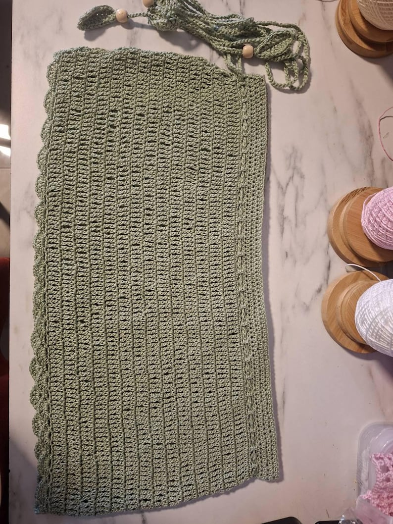 May include: A sage green, rectangular crochet piece with a scalloped edge. The handmade item features a textured stitch pattern and a decorative cord with wooden beads. The piece is likely a handmade home decor item.