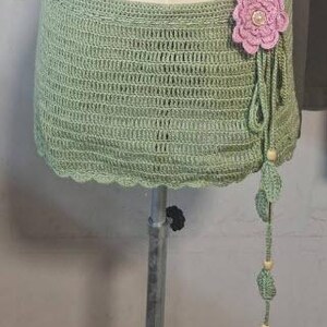 May include: A sage green crochet skirt with a scalloped edge. A large pink flower with a pearl center adorns the side, with dangling green leaves and wooden beads. The skirt is displayed on a mannequin.