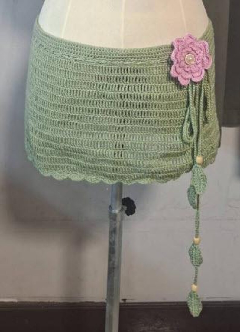 May include: A sage green crochet skirt with a scalloped edge. A large pink flower with a pearl center adorns the side, with dangling green leaves and wooden beads. The skirt is displayed on a mannequin.
