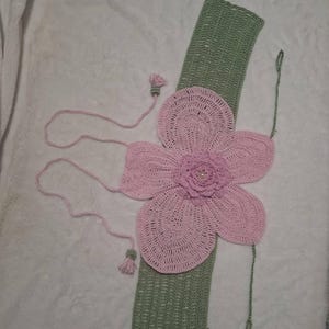 May include: A handmade crochet flower accessory in shades of pink and green. The large pink flower is centered on a green rectangular base. Pink strings and tassels extend from the flower. The accessory is likely for decorative purposes.