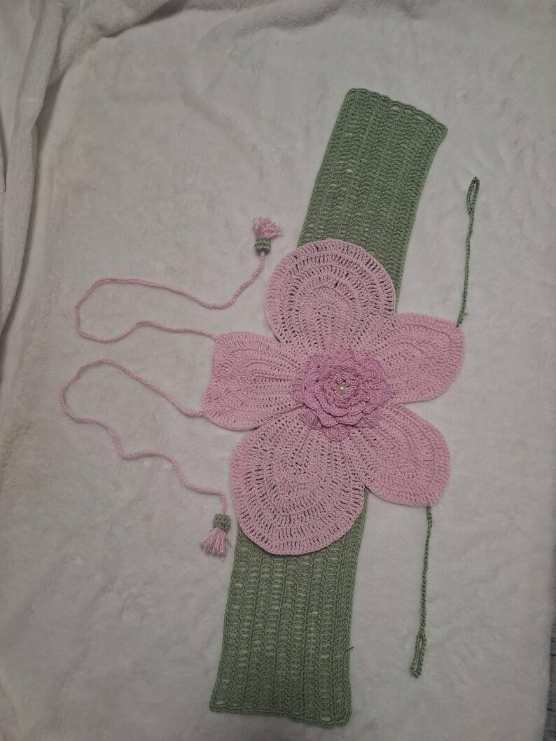 May include: A handmade crochet flower accessory in shades of pink and green. The large pink flower is centered on a green rectangular base. Pink strings and tassels extend from the flower. The accessory is likely for decorative purposes.