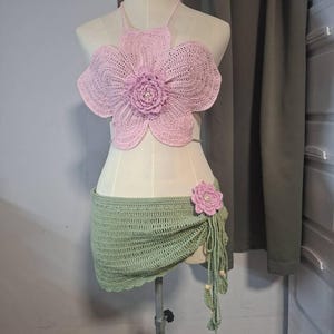 May include: A handmade crochet two-piece set. The top is a light pink flower shape with a pearl center. The skirt is a light green wrap with a matching flower accent and beaded fringe. The set is displayed on a mannequin.