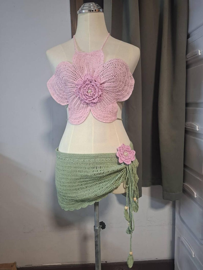 May include: A handmade crochet two-piece set. The top is a light pink flower shape with a pearl center. The skirt is a light green wrap with a matching flower accent and beaded fringe. The set is displayed on a mannequin.