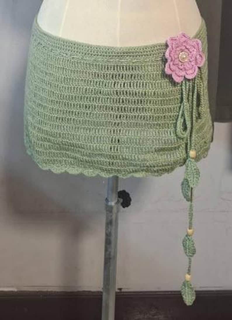 May include: A sage green crochet skirt with a scalloped edge. A large pink flower with a pearl center adorns the side, with dangling green leaves and wooden beads. The skirt is displayed on a mannequin.