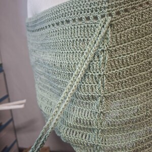 May include: A light green crochet wrap with a textured, open weave design. The wrap features a scalloped edge and long ties for securing. The handmade garment is displayed on a mannequin, showcasing its shape and drape.