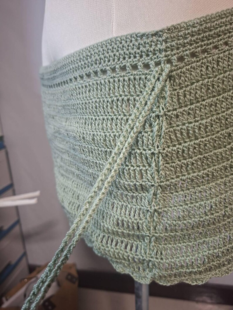 May include: A light green crochet wrap with a textured, open weave design. The wrap features a scalloped edge and long ties for securing. The handmade garment is displayed on a mannequin, showcasing its shape and drape.