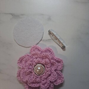 May include: A handmade, light pink crochet flower with a pearl center, a white felt circle, and a safety pin. The flower has multiple layers of petals and is likely a brooch or embellishment for clothing or accessories.