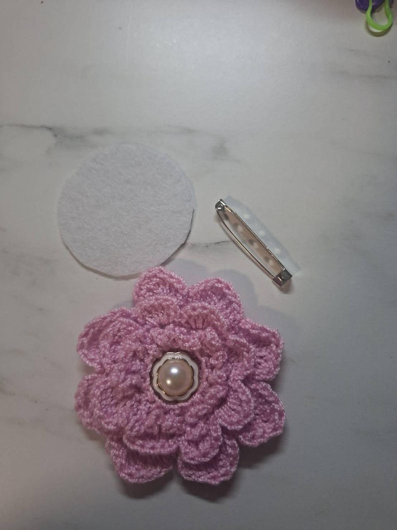 May include: A handmade, light pink crochet flower with a pearl center, a white felt circle, and a safety pin. The flower has multiple layers of petals and is likely a brooch or embellishment for clothing or accessories.