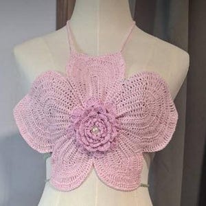 May include: A light pink, handmade crochet top in the shape of a large flower. The top features a halter neck and a central flower design with a pearl bead. The crochet work is visible, showcasing the texture and pattern of the yarn.