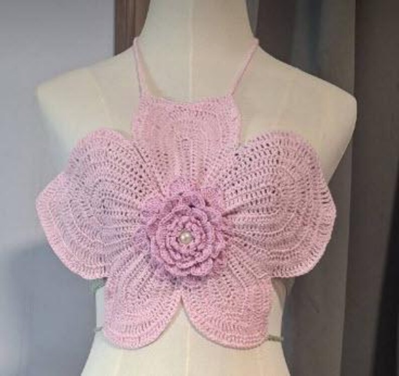 May include: A light pink, handmade crochet top in the shape of a large flower. The top features a halter neck and a central flower design with a pearl bead. The crochet work is visible, showcasing the texture and pattern of the yarn.